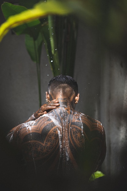 Free photo shirtless muscular latin american male with tattoos standing taking shower outloodrs after workout on the tropical leaves background. shower in the private villa.