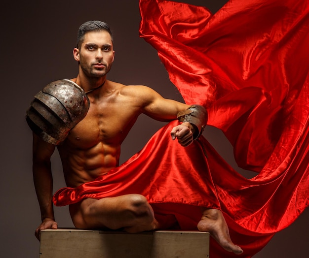 Free photo shirtless muscular man sitting on a podium. red cloth on grey background