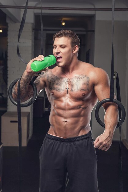 Shirtless muscular man with tattoo relaxing after ring workouts in a gym club.