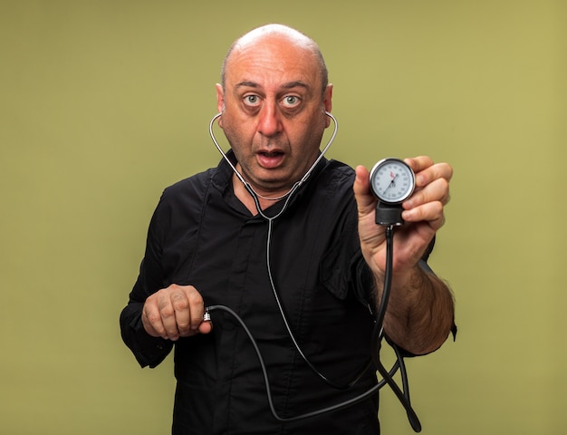 shocked adult ill caucasian man holding sphygmomanometer isolated on olive green wall with copy space