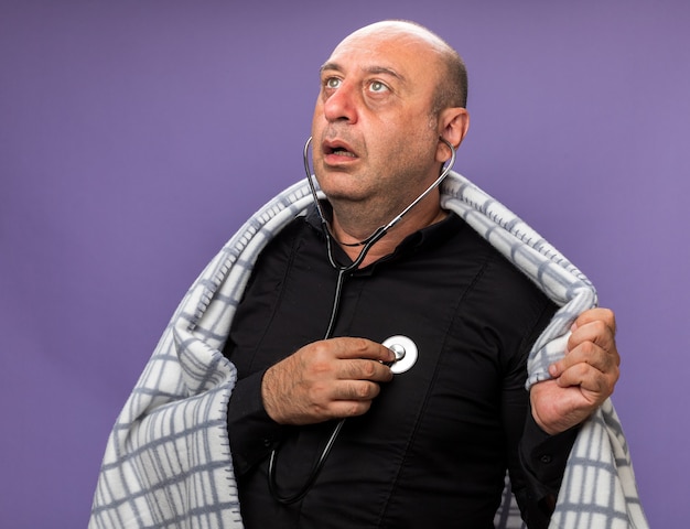 shocked adult ill caucasian man wrapped in plaid measuring heart beat with stethoscope looking at side isolated on purple wall with copy space