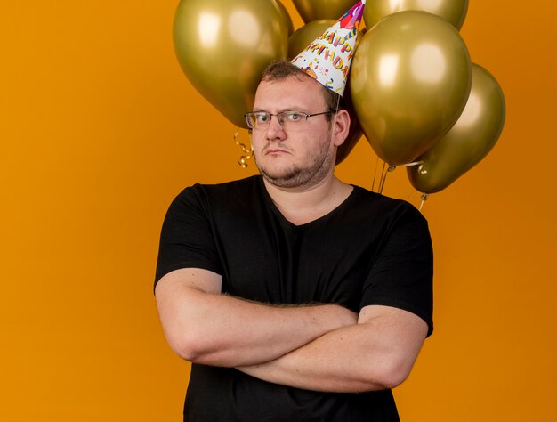 Shocked adult slavic man in optical glasses wearing birthday cap stands with crossed arms in front of helium balloons 