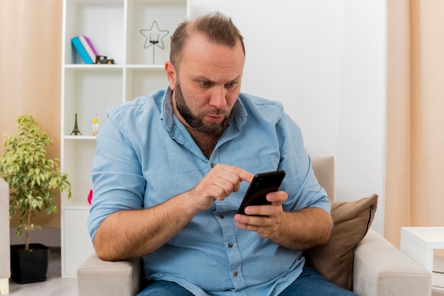 Shocked adult slavic man sits on armchair pointing and looking at phone inside the living room