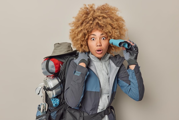 Free photo shocked amazed female tourist notices something breathtaking into distance holds binocular wears jacket and gloves carries big rucksack with necessary equipment isolated over grey background