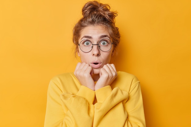 Shocked amazed young European woman gasps and keeps hands on cheeks stares surprised keeps mouth opened wears round spectacles for sight and casual jumper isolated over vivid yellow background