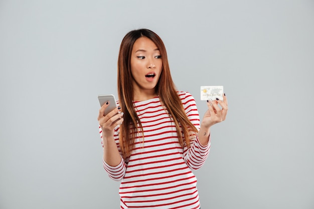 Free Photo shocked asian woman in sweater using smartphone and credit card over gray background