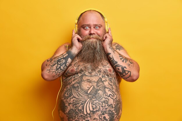 Shocked bald man with naked obese body, has tattooed arms and belly, thick beard, chills with good vibes, listens music in headphones, enjoys great sound, isolated on yellow wall.