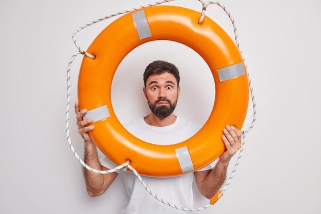 Free Photo shocked bearded adult man looks through inflated lifebuoy stares bugged eyes learns how to swim or rescue people wears casual t shirt isolated over white background male lifesaver poses indoor