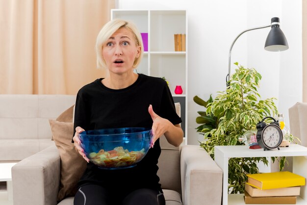 Shocked beautiful blonde russian woman sits on armchair holding bowl of chips looking forward inside living room