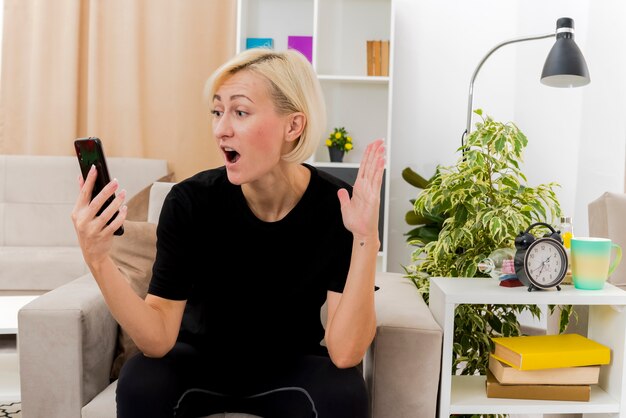 Shocked beautiful blonde russian woman sits on armchair raising hand and looking at phone inside the living room