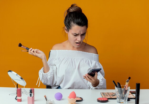 Shocked beautiful girl sits at table with makeup tools holds makeup brushes looking at phone isolated on orange wall