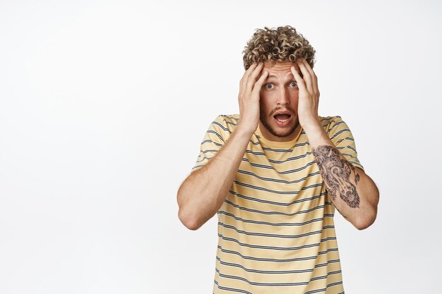 Shocked blond guy hold his head in panic looking anxious and alarmed scared of something terrible happening standing over white background