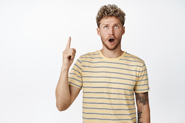 Shocked blond man drop jaw while looking and pointing up showing shocking sale discounts in shop standing in tshirt over white background