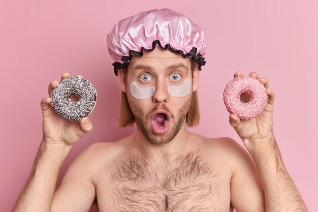 Shocked blue eyed adult guy stares keeps mouth opened applies patches under eyes for skin treatment wears bath hat holds two glazed donuts stands shirtless