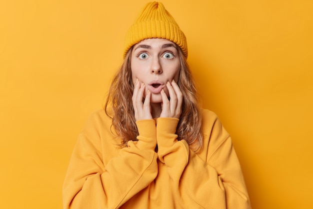 Shocked blue eyed young woman stares at camera keeps hands on cheeks mouth widely opened wears casual sweatshirt and hat reacts on something scarying isolated over yellow background Omg concept