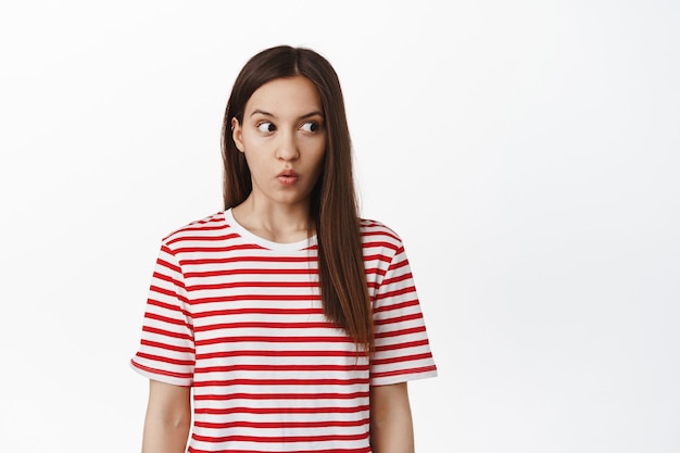 Shocked brunette woman gasping, look right at empty space with speechless startled expression, checking out smth impressive, standing against white wall.