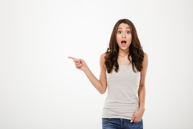 Shocked brunette woman with arm in pocket pointing on copyspace and looking at the camera over gray