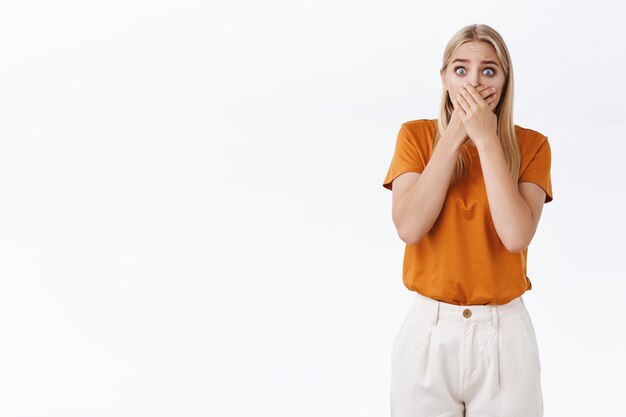 Shocked concerned and frightened young blond woman with tattoos, gasping cover mouth with palms and stare camera scared, freak-out from stunning news, standing white background