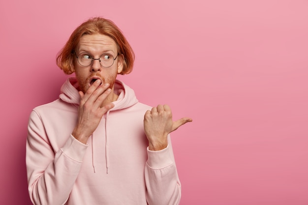 Shocked concerned ginger hipster covers opened mouth, expresses amazement, shows something disturbing, stands astounded, points thumb aside, tells you urgent shocking news, wears sweatshirt.