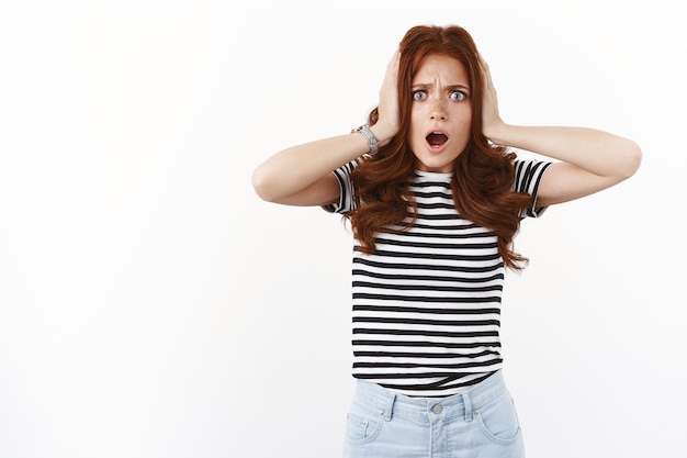 Shocked concerned young ambushed redhead woman in striped t-shirt, hold hands on head, furrow eyebrows nervously, open mouth ambushed and worried, standing disappointed and distressed