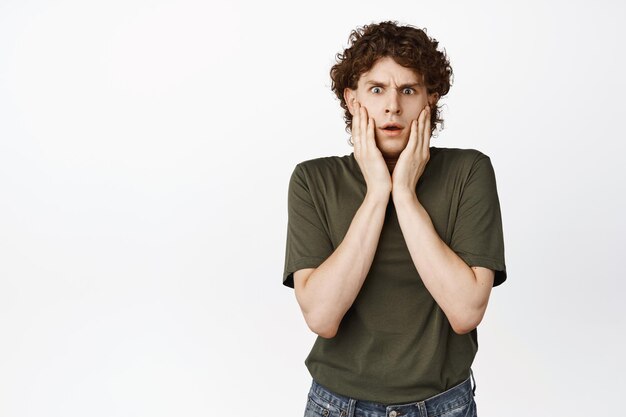 Shocked and concerned young funny guy staring confused at camera touching his face in disbelief standing over white background copy space