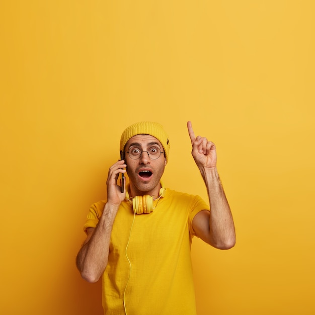 Free photo shocked concerned young man surprised with troublesome news, points fore finger above, wears spectacles and yellow outfit