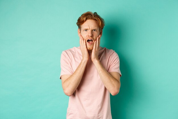 Shocked and concerned young man with red hair, staring at camera worried and touching face, standing in t-shirt against turquoise background.