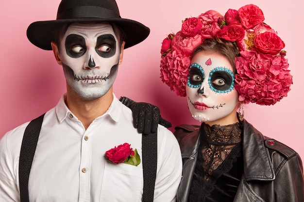 Shocked couple have scary face expressions, funky makeup and costumes, wear black and white attire decorated with red flowers, pose together in studio against pink wall