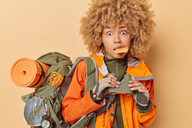 Shocked curly haired woman spends vacation actively keeps waffle in mouth holds binoculars carries heavy rucksack dressed in casual clothes isolated over brown background Tourist adventure