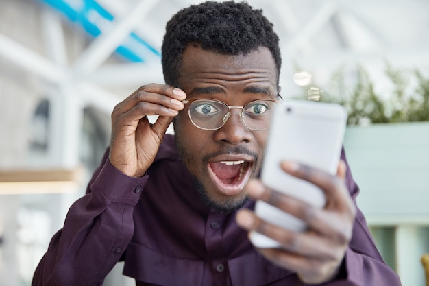 Shocked dark skinned young man stares with bugged eyes, keeps jaw dropped, wears transparent glasses