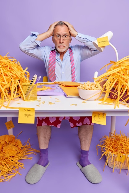 Free photo shocked embarrassed elederly grey haired man keeps hands on head poses at messy desktop doesnt know from what to start working wears formal shirt and tie around neck works at home omg concept