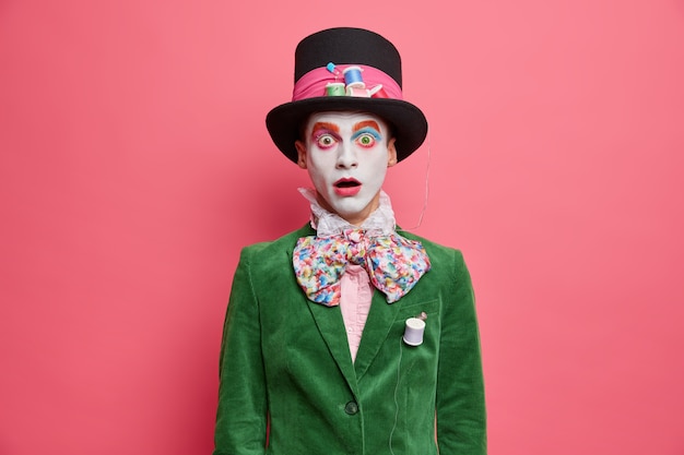 Free photo shocked embarrassed hatter stares at camera with bugged eyes dressed in green jacket bowtie and big hat has colorful makeup takes part in performance isolated over rosy wall