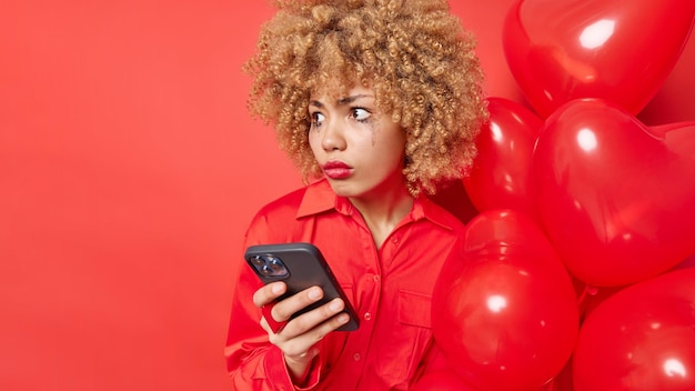 Shocked embarrassed woman looks away with stunned expression sees something amazing and breathtaking uses smartphone for online communication poses with bunch of balloons against red studio wall