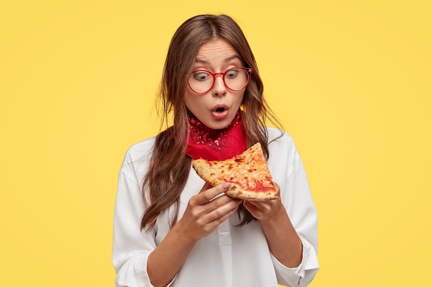 Free photo shocked emotive lady looks with surprisement at slice of pizza, amazed with its wonderful taste, wears spectalces and white shirt, models against yellow wall. people, reaction and nutrition concept