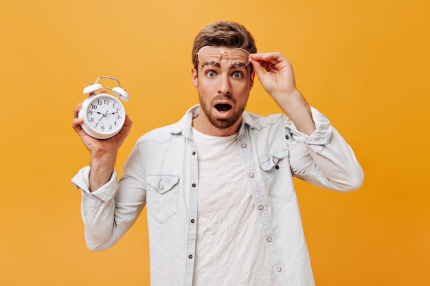 Shocked fashionable guy with blue eyes and ginger beard in white outfit taking off eyeglasses looking into camera and holding clock