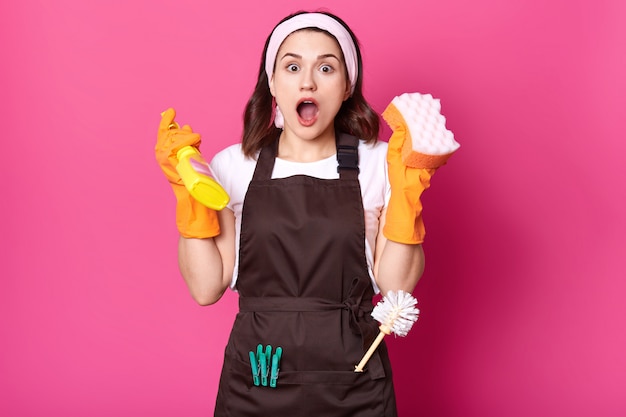 Free Photo shocked female housekeeper holds sponge and detergent spray in hands, having much work to do. attractive woman with surprised and excited look wearing apron and protective gloves. copy space.