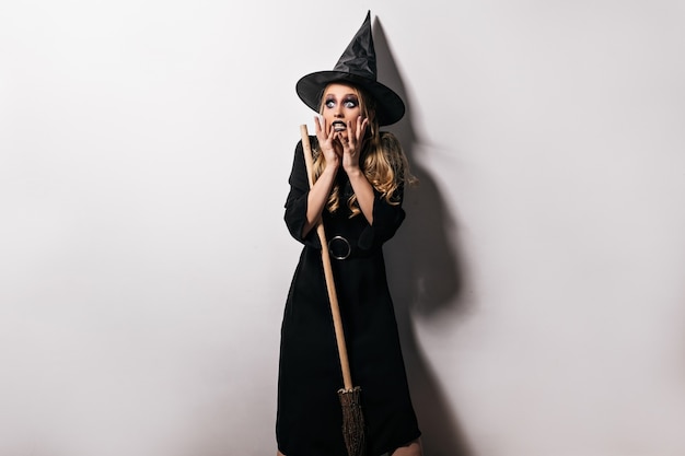 Free Photo shocked female model in wizard costume posing on white wall. indoor shot of amazed witch girl standing with scared face expression.