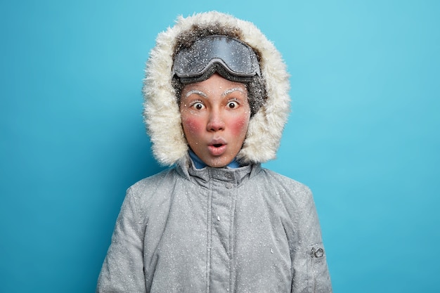 Free photo shocked female skier with red frozen face stares dressed in grey jacket with hood and snowboarding goggles.