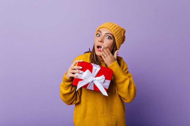 Free photo shocked girl in casual sweater and hat posing with gift. indoor portrait of debonair lady with new year present expressing amazement.