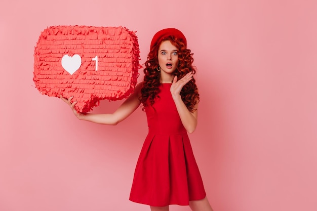 Shocked girl looks at camera holding like sign on pink background Portrait of woman in red dress and beret