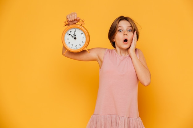 Shocked girl showing alarm clock and looking camera with opened mouth isolated