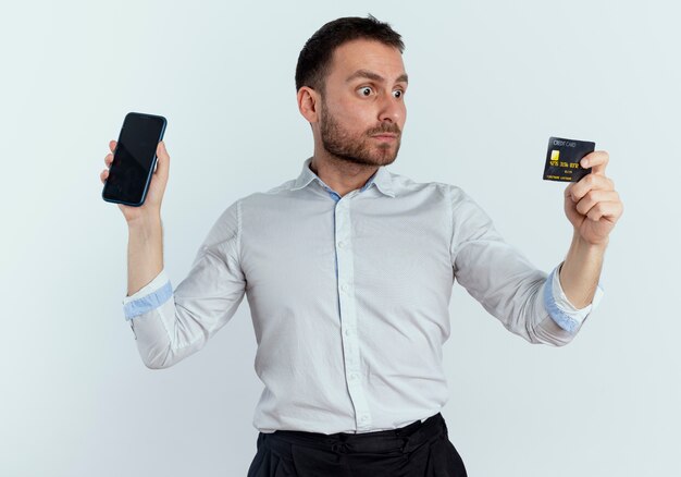 Shocked handsome man holds phone and looks at credit card isolated on white wall