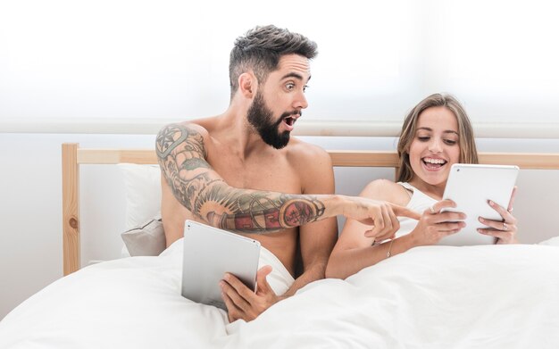 Shocked man looking at his happy wife using digital tablet on bed