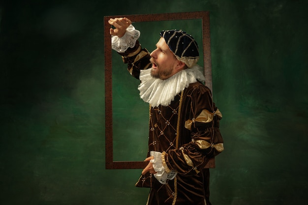 Free photo shocked. portrait of medieval young man in vintage clothing with wooden frame on dark background. male model as a duke, prince, royal person. concept of comparison of eras, modern, fashion, sales.