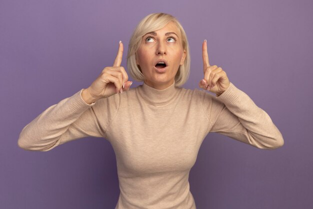 Shocked pretty blonde slavic woman looks and points up with two hands isolated on purple wall