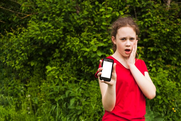 Shocked pretty girl showing blank screen cellphone while standing at park