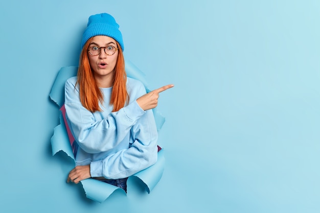 Free photo shocked red haired young woman holds breath stares bugged eyes indicates away on copy space stands in ripped hole wears blue hat and jumper demonstrates something of high price on item