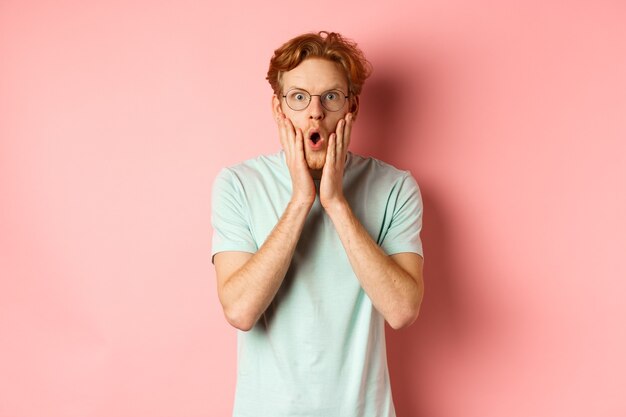 Shocked redhead man gossiping staring impressed at camera and touching face standing in glasses agai...