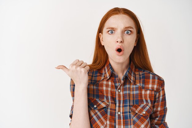 Shocked redhead woman with pale skin and no makeup pointing finger left at banner gasping amazed at camera standing over white background