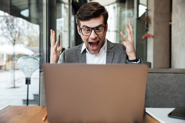 Shocked screaming businessman in eyeglasses sitting by the table in cafe and rejoice while looking at laptop computer